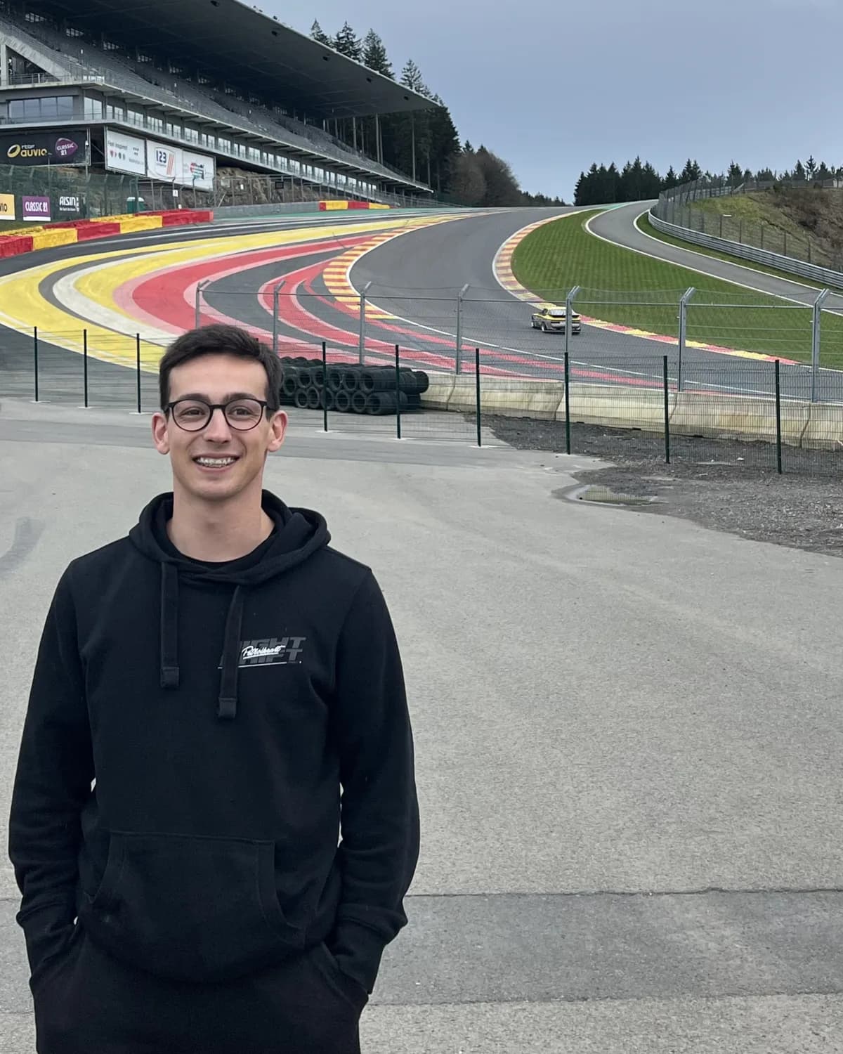 João Paulo Santos, smiling in a black hoodie, standing trackside at Spa-Francorchamps with the Raidillon climb and the main grandstand behind him.